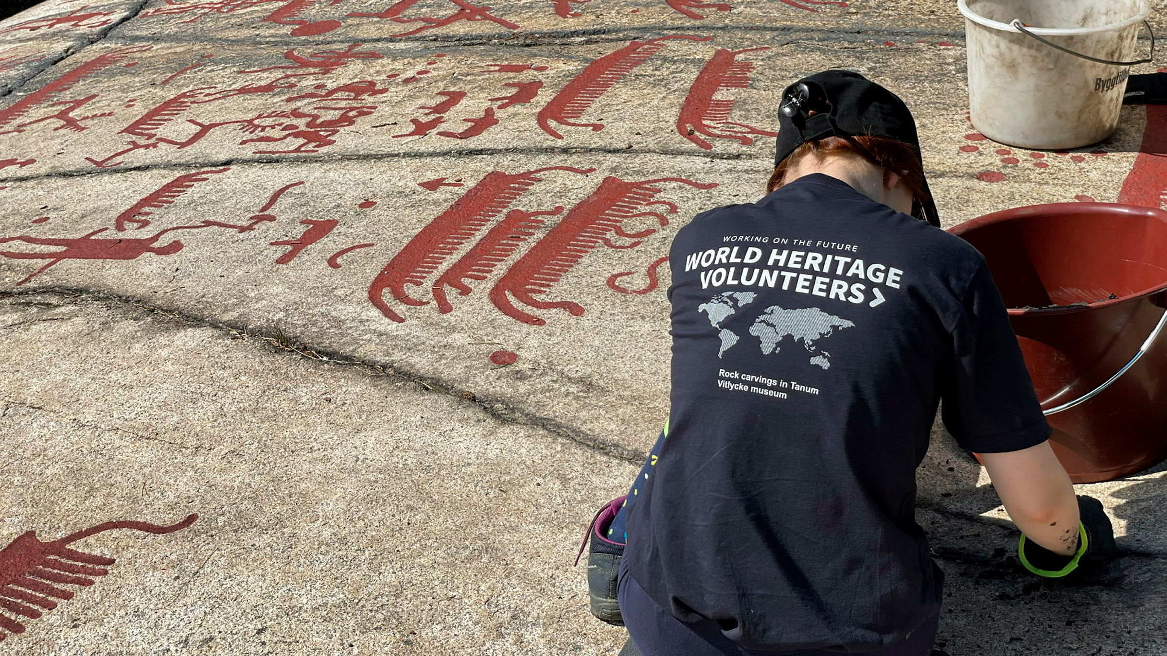 En person i mörk t-shirt, keps, byxor och handskar sitter med ryggen mot kameran vid en hällristning och rengör den.