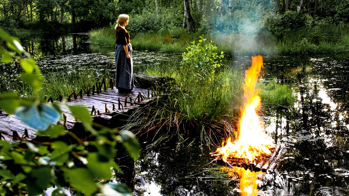 Bild på kvinna i bronsålderskläder och en flotte med en eld på.