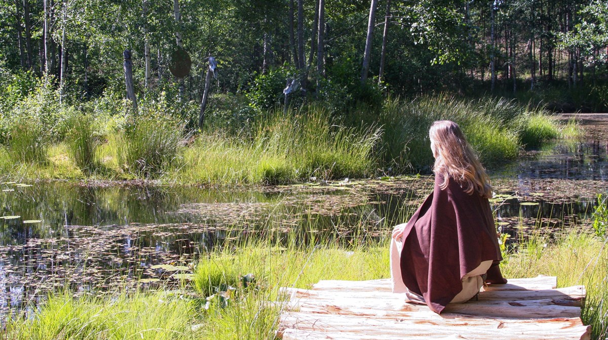 En kvinna i ullkläder knäböjer framför en gräsomgärdad vattenspegel