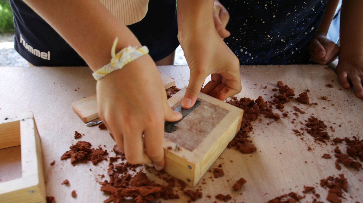 Två händer pressar ner en bronsrakkniv i en av gjutsand uppfylld träram. Gjutsanden är brunröd och ytan är täckt av ett vitt pulver.