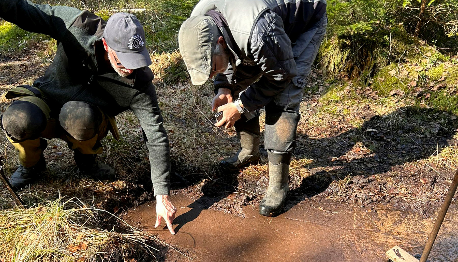 Två män i heltäckande kläder och kepsar står vid en hällristning, Den ena pekar mot en hällristning.